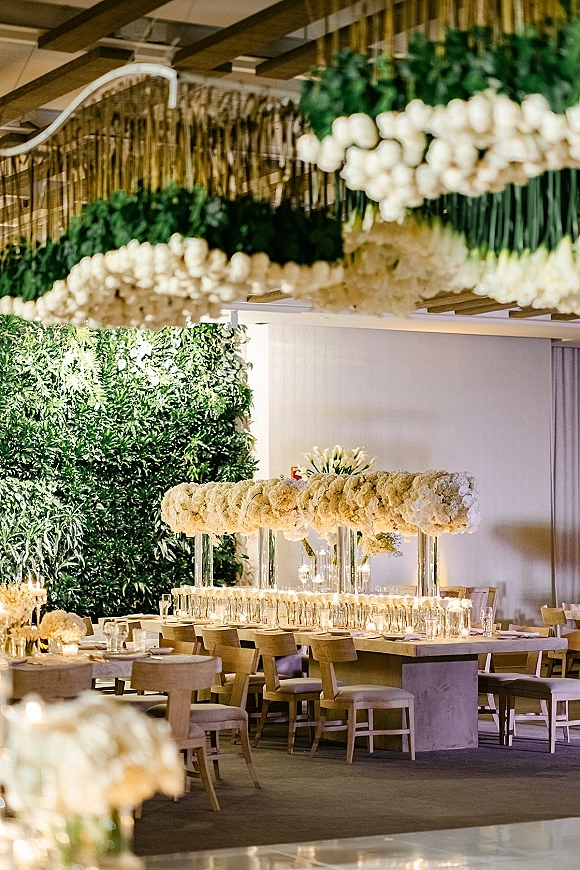 Reception tablescape with long banquet table wedding styling, white floral runner and candlelit glass votives beneath hanging florals and greenery wall backdrop