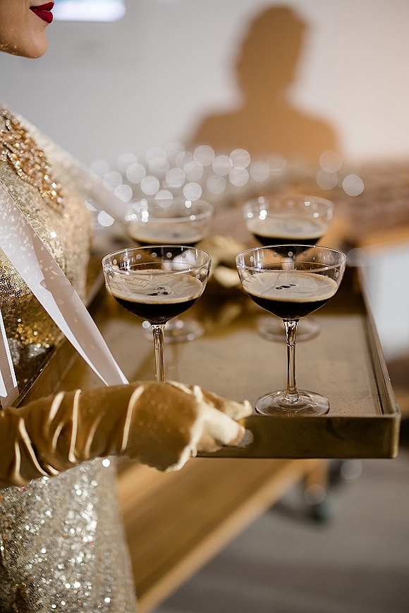 Champagne coupe toast on a serving tray, held by a server in a metallic sequin dress and long gloves, with bokeh-lit guests behind