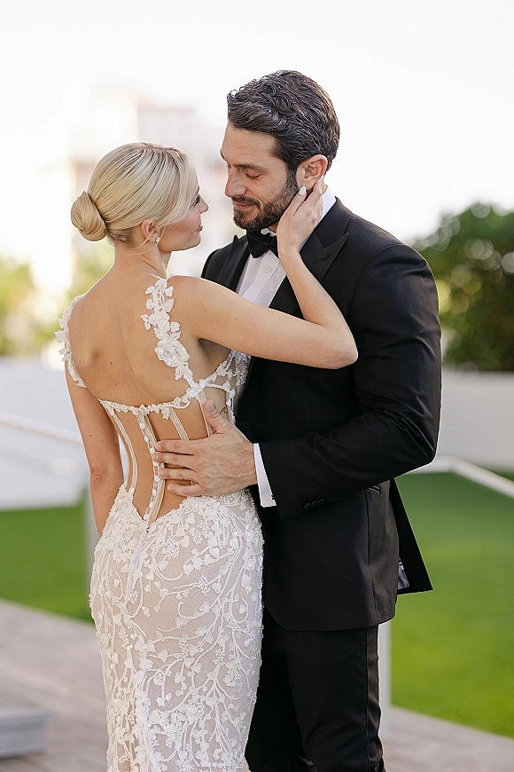 Couple portrait of bride and groom embrace, bride touching groom’s face in lace gown on a terrace with white railing and trees