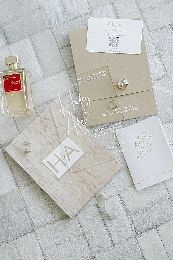 Wedding invitation suite with acrylic wedding invitation, QR code RSVP, rings, vow book and perfume arranged on a white quilted bedspread
