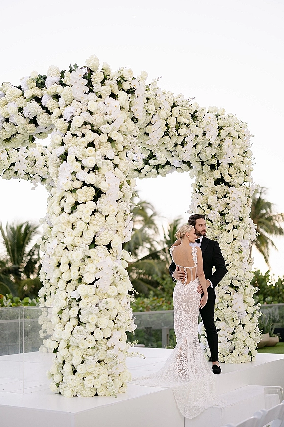 Couple portrait of bride and groom posing in lace wedding dress and black tuxedo beneath a white floral arch on a palm-lined terrace