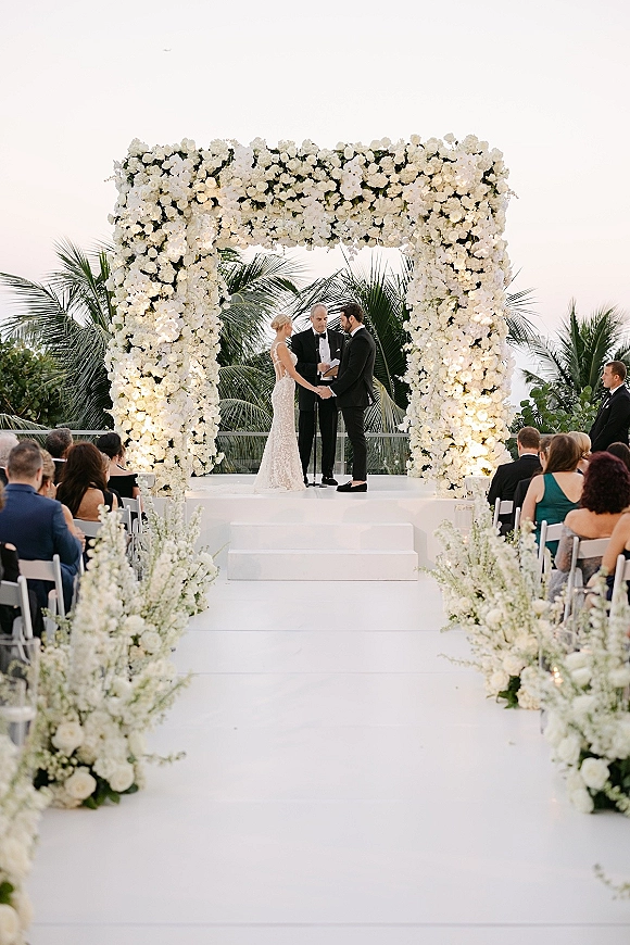 Wedding ceremony with couple exchanging vows under a white floral arch, candles and aisle florals on a white runner, palm trees behind guests