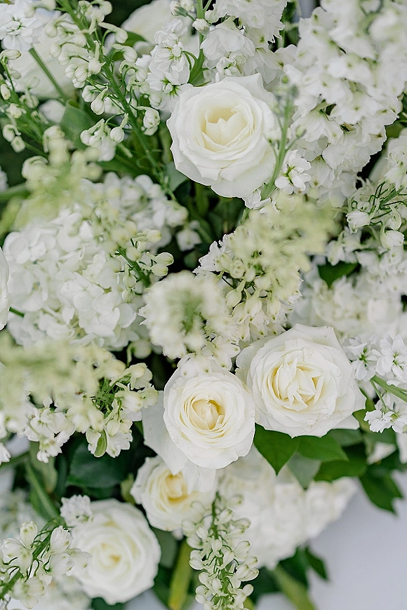 Wedding floral arrangement of white roses and airy filler blooms with greenery, stems visible against softly blurred florals background