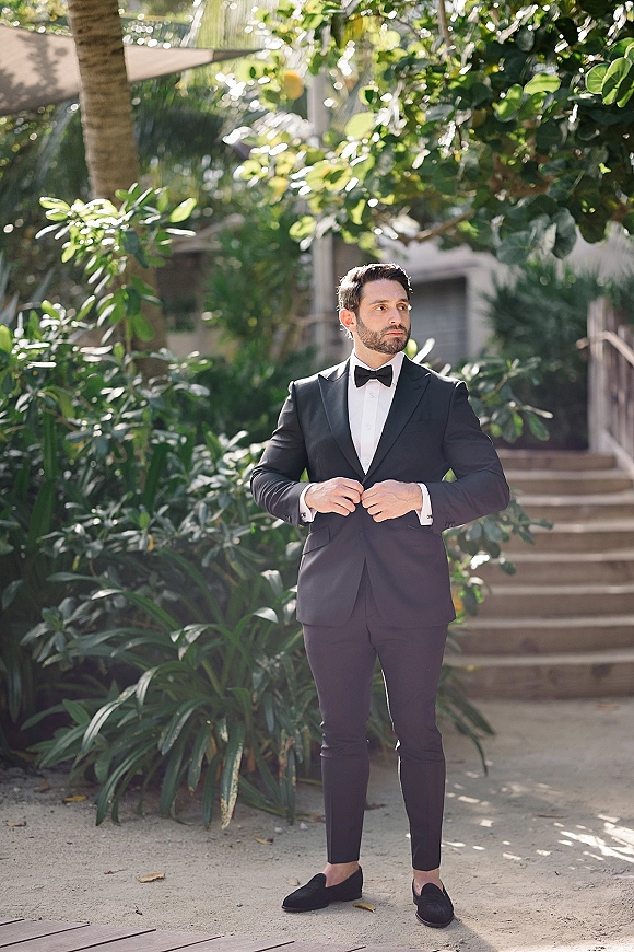 Groom portrait in a black tuxedo, bow tie and cufflinks, standing on garden stairs with lush greenery and sunlit building behind