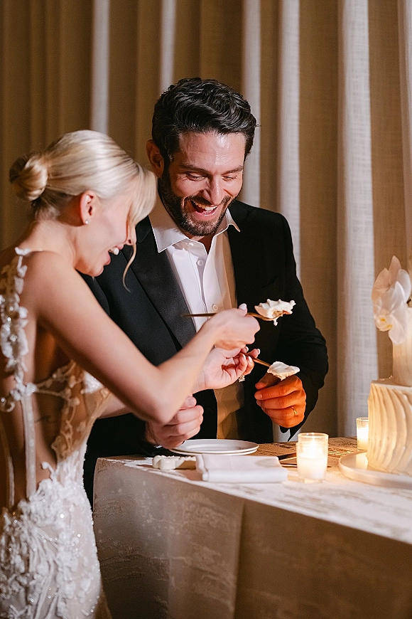 Cake cutting moment with bride and groom slicing a white buttercream wedding cake at a candlelit table with draped curtains behind