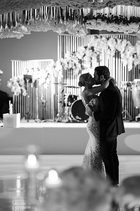 Wedding kiss during first dance as bride in strapless lace gown embraces groom in black suit beneath hanging floral ceiling in ballroom
