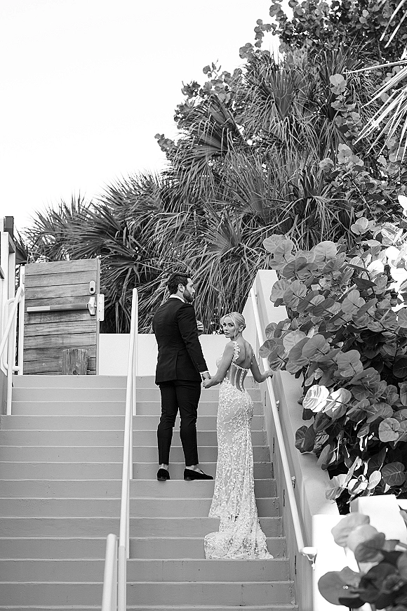 Couple portrait of bride and groom holding hands on an outdoor staircase, bride looking back in a lace dress train amid tropical foliage