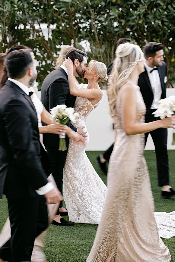 Wedding kiss as the bride and groom kiss in front of their wedding party, lace dress and bouquets against a green lawn and hedge backdrop
