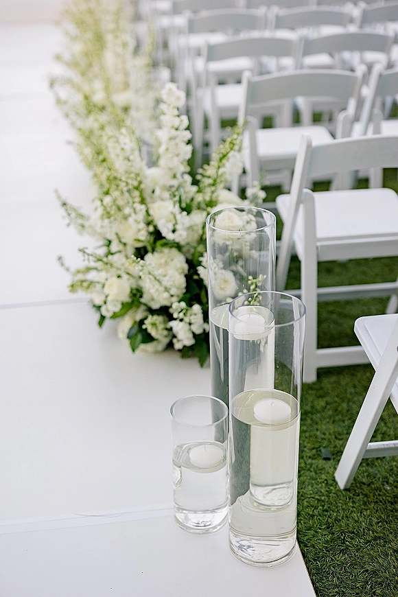 Ceremony aisle decor with a white aisle runner, white floral arrangements and greenery in glass cylinder vases with floating candles on a lawn