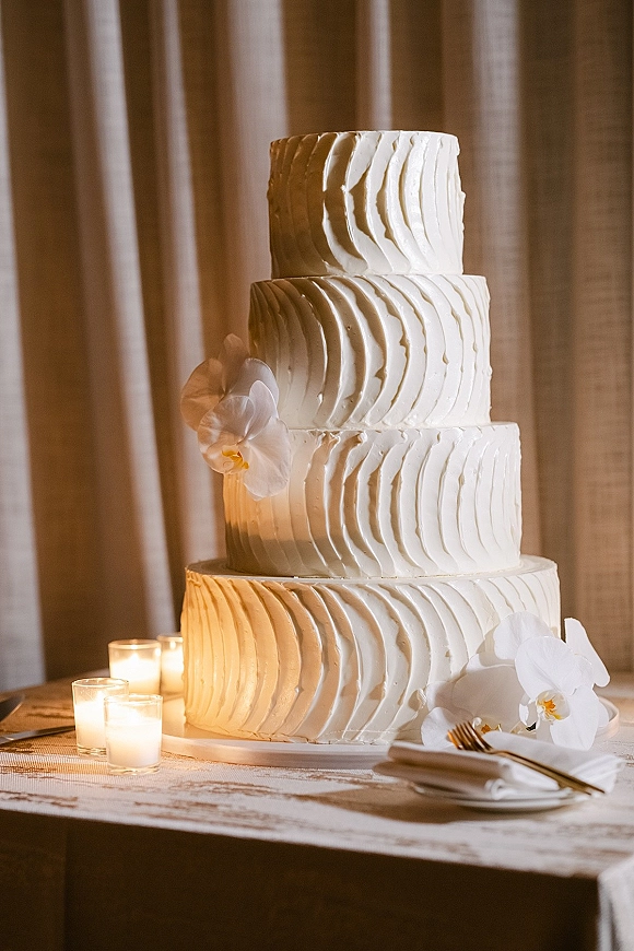 Wedding cake with textured buttercream frosting, orchid accents, and votive candles on a stand, set on a table before draped curtains