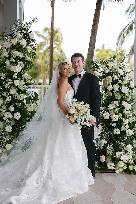 Couple portrait of bride and groom smiling under a white rose and greenery arch, her strapless lace gown and long veil on a palm-lined terrace