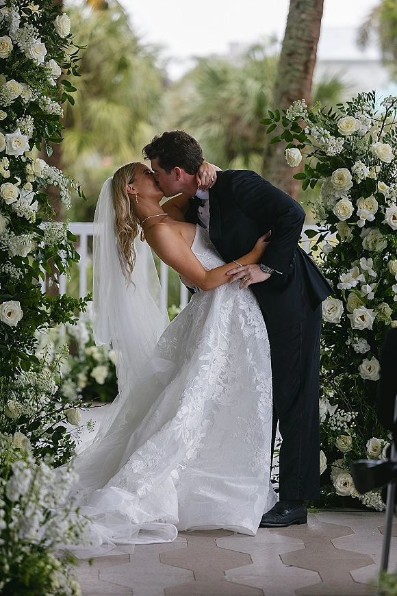 Wedding kiss as the groom dips the bride beneath a white rose and greenery floral arch, veil flowing in an outdoor garden with palm trees