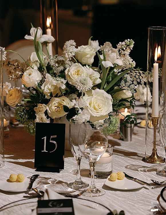 Reception tablescape with a wedding table centerpiece of white roses, taper candles and glass hurricanes on a white linen table in dim light