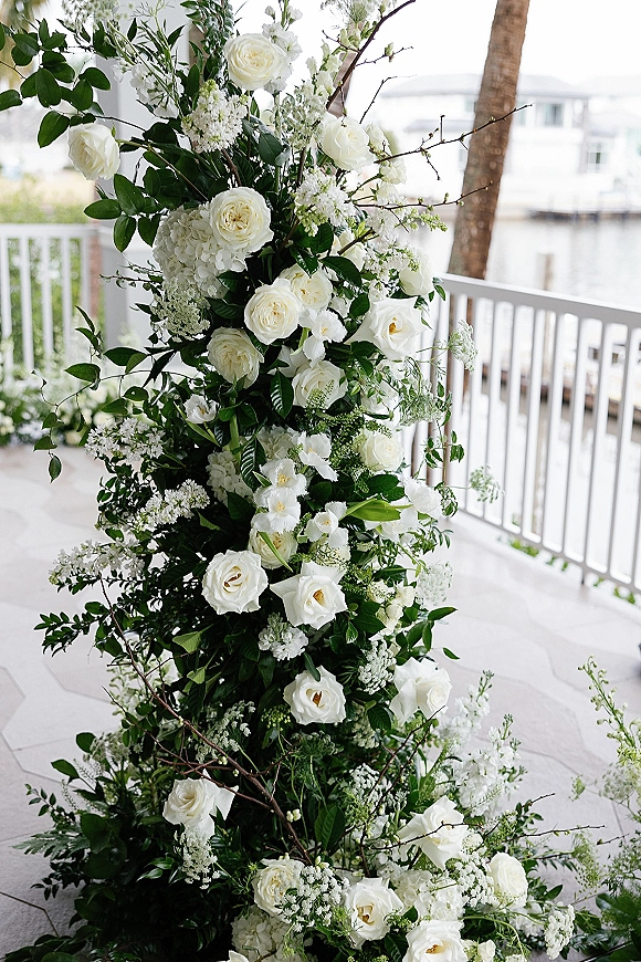 Wedding floral installation with white roses accents and hydrangea blooms, greenery and branches styled by a veranda railing with water view