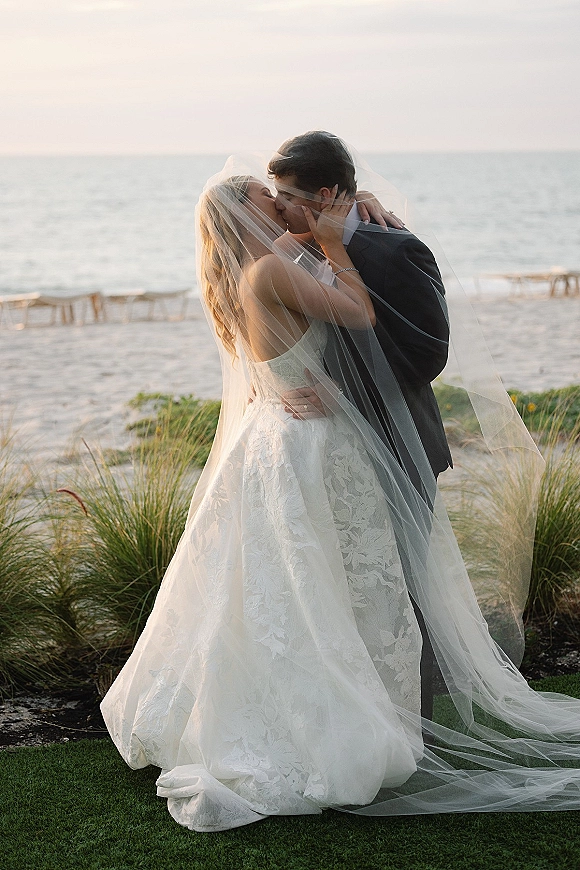 Wedding kiss portrait of bride and groom kissing under her veil, lace gown and black suit by an ocean shoreline at golden hour