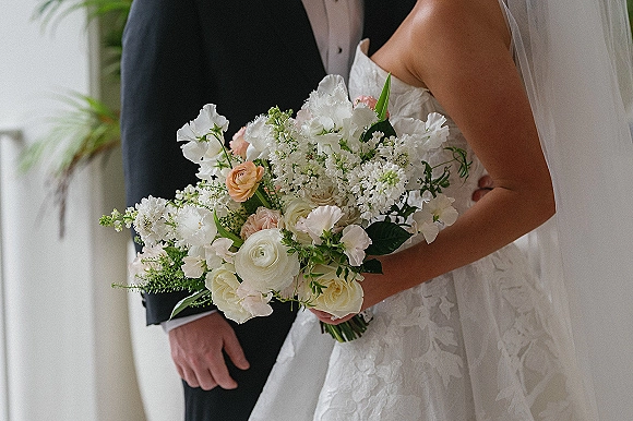 Bridal bouquet of blush and white flowers with greenery held by a bride in a lace gown and veil beside a groom in tux near a window