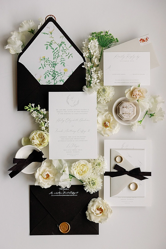 Invitation suite with black and white wedding invitations in black envelopes, botanical liner, wax seal, ribbon, and gold bands on neutral surface