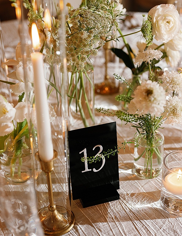 Reception tablescape wedding table centerpiece with white flowers in bud vases, taper candles in brass holders, and a black table number card under warm light