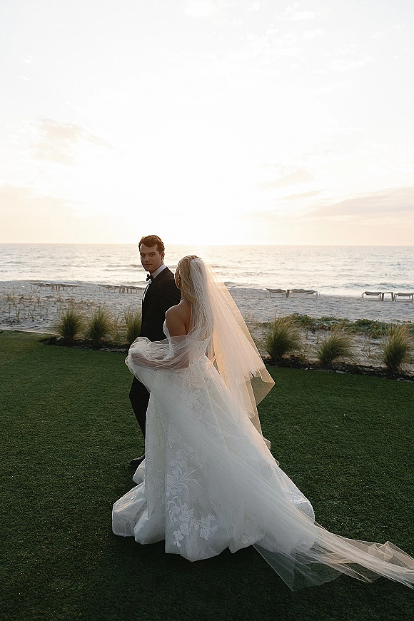 Couple portrait on a beach wedding portrait at sunset, bride in strapless lace gown with long veil and groom in tuxedo by the ocean shoreline