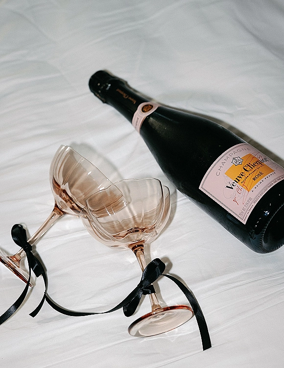 Champagne bottle for a wedding champagne toast with pink coupe glasses tied in black bows on white bed sheets flat lay