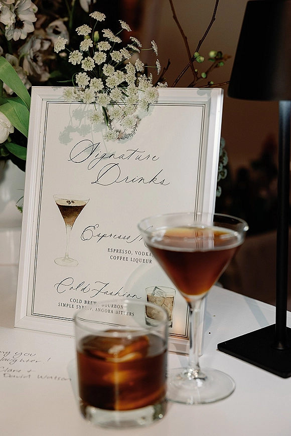 Signature drinks sign with calligraphy cocktail menu in a framed display, featuring martini and rocks glass illustrations on a reception table