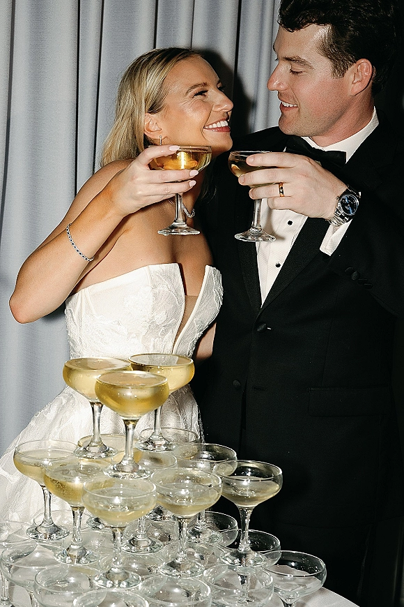 Wedding toast as bride in a strapless gown and groom in a black tuxedo clink champagne coupe glasses beside a champagne tower backdrop