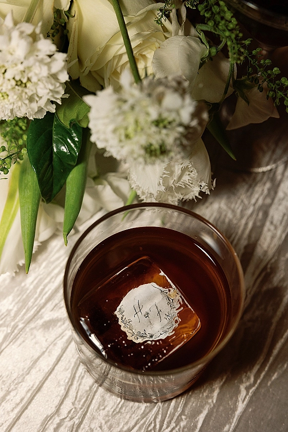 Signature cocktail wedding signature drink in a rocks glass with monogrammed ice cube, amber spirit, white roses and hydrangea on linen tabletop