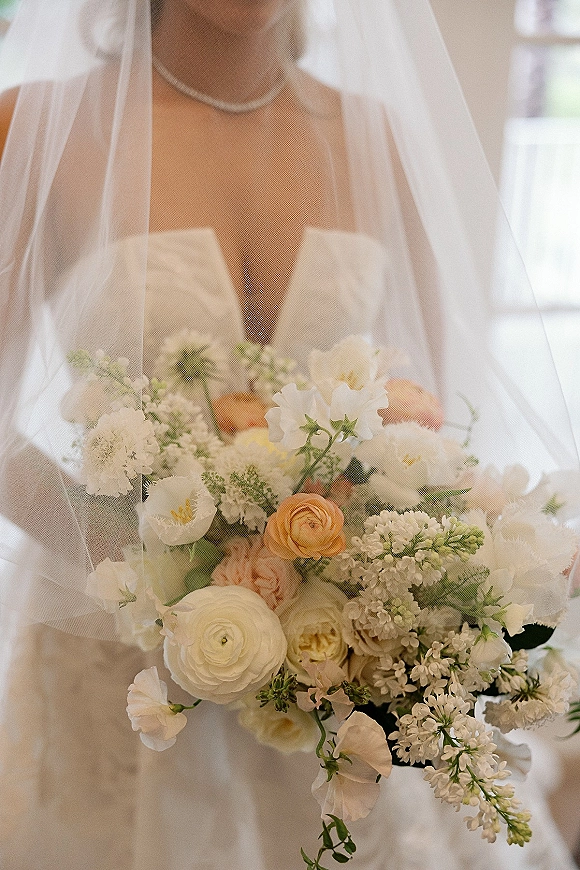 Bridal bouquet of white and peach bouquet blooms with greenery, held against a strapless dress with veil in bright window light indoors