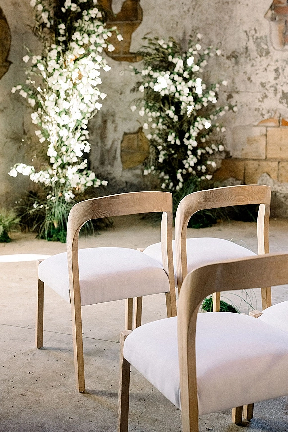 Ceremony seating with wedding ceremony chairs in natural wood, white cushions, and greenery with white florals against plaster and brick wall