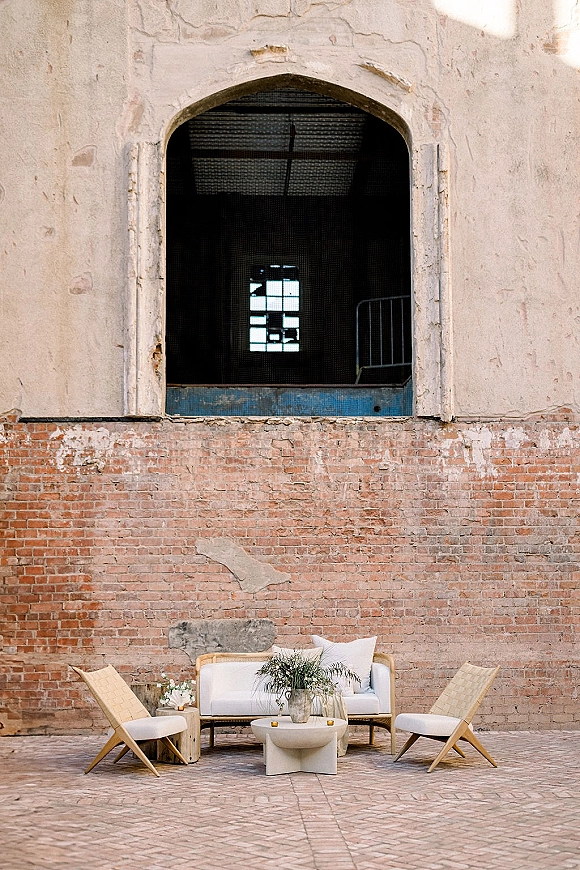 Lounge seating area with wedding lounge furniture, white sofa and wicker chairs, throw pillows, candles and florals against exposed brick wall