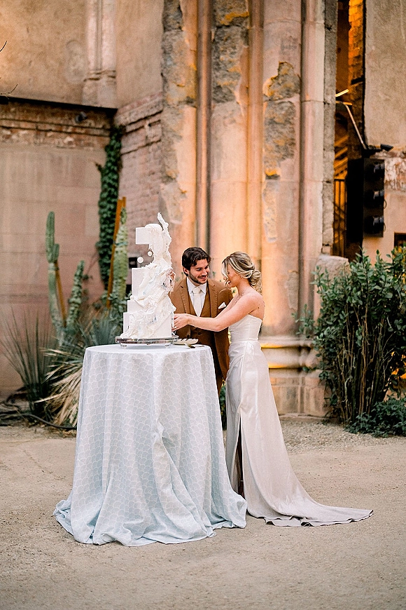 Wedding cake cutting with bride and groom slicing a textured white cake on an acrylic stand outside a stone facade with uplighting and cactus backdrop