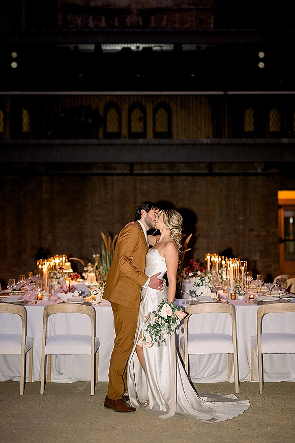 Wedding kiss portrait of bride and groom kiss at a candlelit reception table, bride holding a bouquet in a strapless dress, rustic hall backdrop