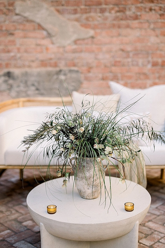 Reception lounge decor with wedding lounge seating, a white sofa and round coffee table topped with stone vase florals and tealight candles against exposed brick