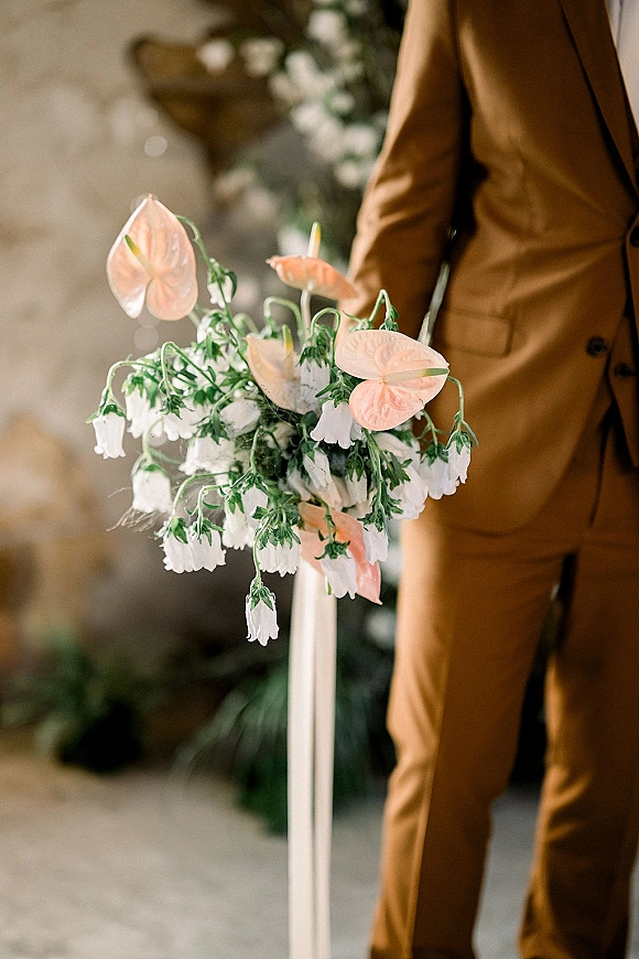 Wedding bouquet with anthurium wedding bouquet blooms, white bell flowers and greenery held by groom in suit before stone wall and pedestal