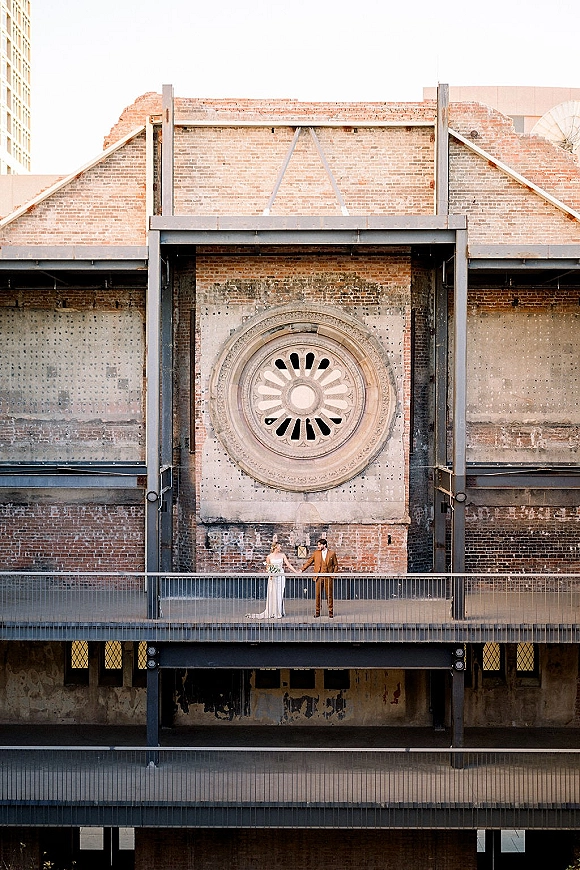 Couple portrait of bride and groom holding hands, her strapless gown and bouquet by a brick wall balcony with round window backdrop