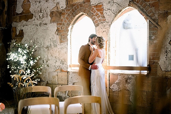 Wedding kiss portrait of bride and groom kissing by arched windows, her hand on his face, backlit against brick wall indoors