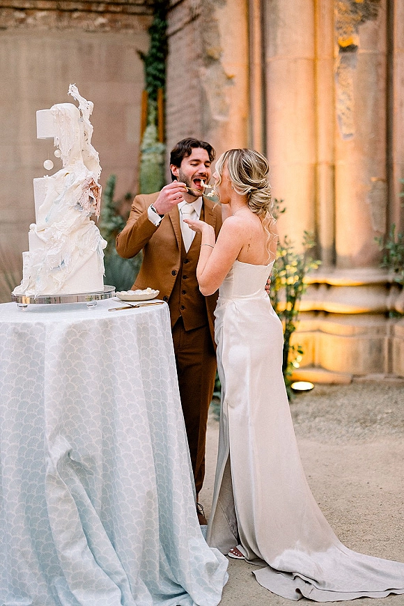 Wedding cake cutting as bride in a strapless satin dress and groom in a brown suit slice a textured cake in an outdoor courtyard setting