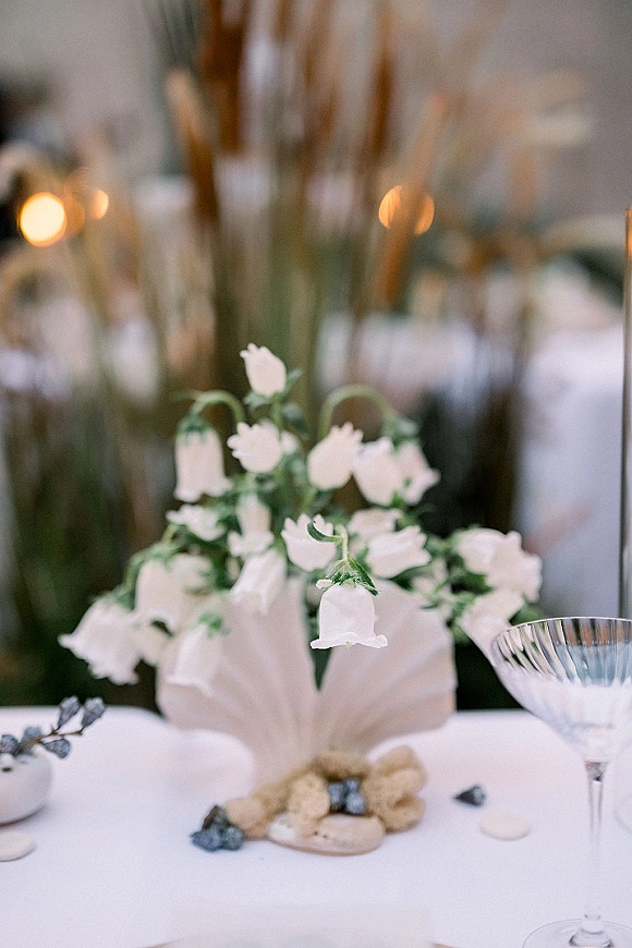 Reception tablescape with a wedding table centerpiece of white flowers in a bud vase, driftwood and pebbles on linen, bokeh lights behind