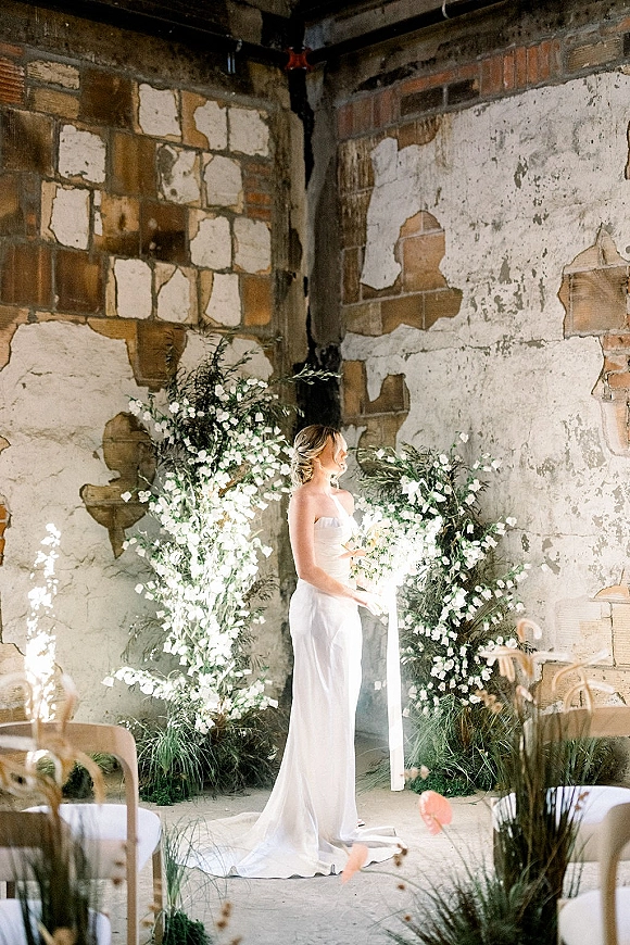 Bridal portrait of a bride in a strapless wedding dress beneath a white flower and greenery arch, with aisle candles by an industrial brick wall
