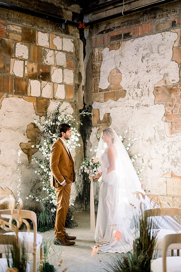 Wedding vows as bride and groom exchange vows in an indoor wedding ceremony, her veil flowing, holding a bouquet before brick walls
