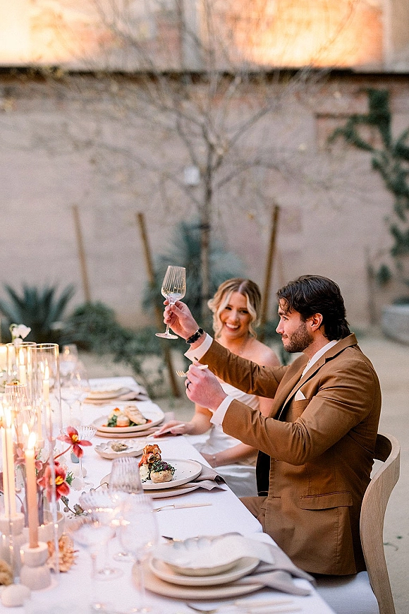 Wedding toast as bride and groom raise champagne flutes at a candlelit al fresco head table in a sunset courtyard with desert plants