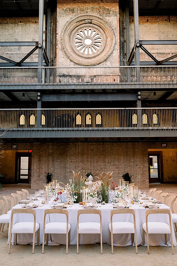 Reception tablescape with long wedding reception table set in white linens, candles, glass cylinder vases and greenery against an industrial brick wall