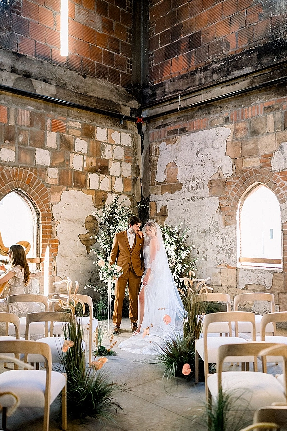 Ceremony moment with bride and groom at altar beneath a floral ceremony arch, her veil flowing in an indoor brick-walled venue