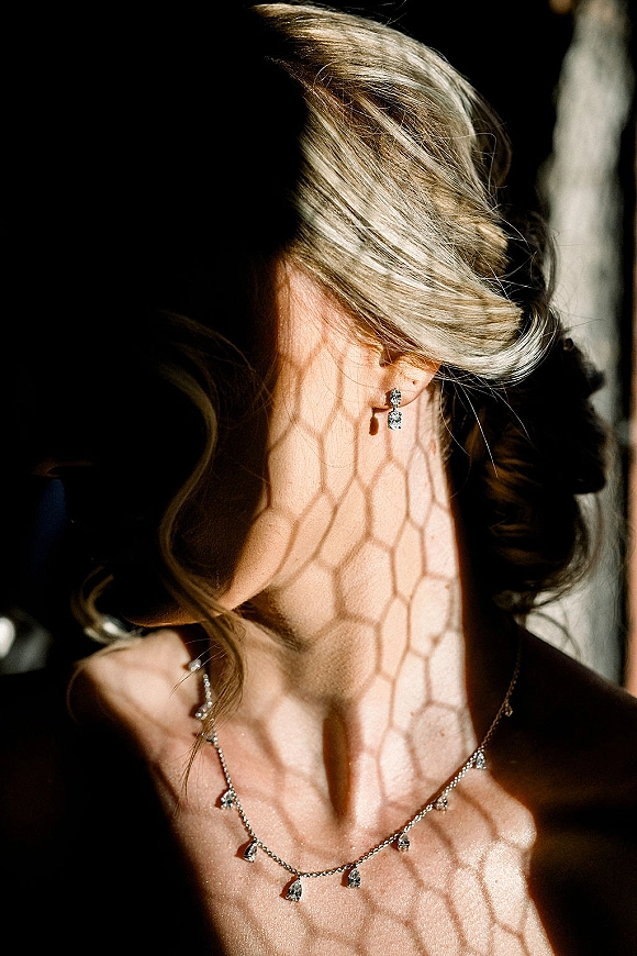 Bridal jewelry with wedding earrings close up, diamond drop earrings and necklace beside an updo, lit by window light in a dark interior