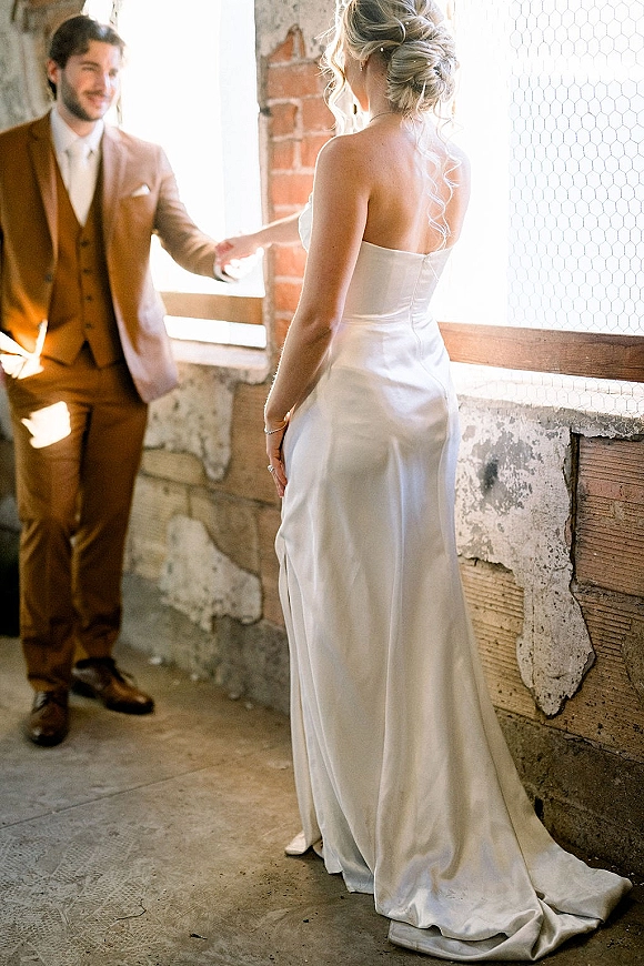 First look moment as bride in strapless satin wedding dress walks toward groom in an industrial loft with window light and exposed brick backdrop
