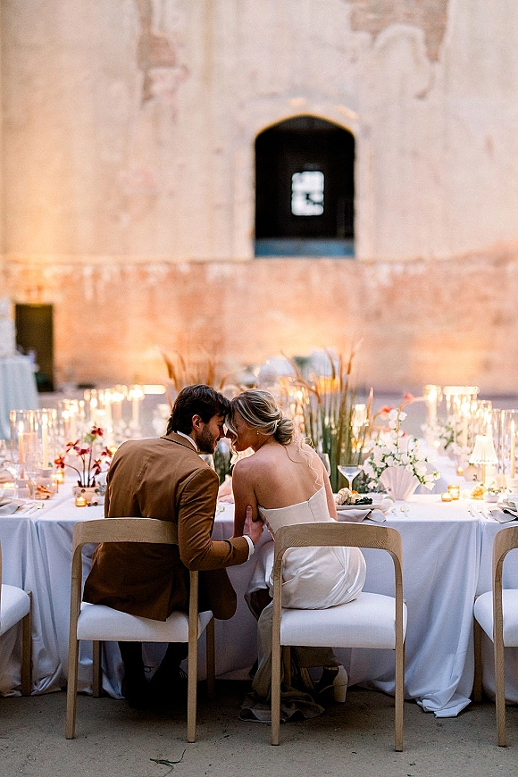 Couple portrait of bride and groom seated holding hands at a candlelit banquet table with taper candles in a stone courtyard with an arched window