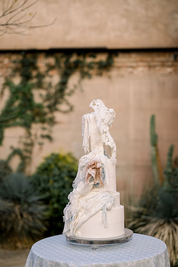 Wedding cake with modern wedding cake styling, white tiered textured icing on a stand atop a lace tablecloth in an outdoor courtyard with cactus greenery