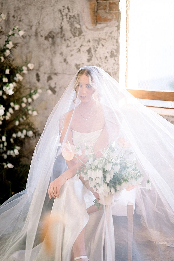 Bridal portrait of a bride with veil sitting by a large window, holding a bouquet with peach tulips against a rustic plaster wall