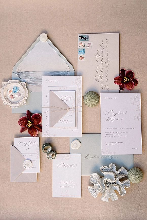 Invitation suite flat lay with calligraphy lettering, wax-sealed envelopes, stamps, RSVP and menu cards, rings, coral and shells on linen backdrop