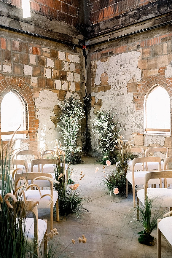 Ceremony setup with white chairs lining an indoor ceremony aisle, grounded florals and pampas grass in natural light by arched brick windows